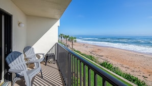 A balcony with two Adirondack chairs, a small table, and a view of a beach with palm trees and the ocean. - Oceanfront Charmer (Ormond Beach)