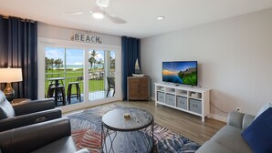 Sala de estar con televisor, ventilador de techo, vista a la playa y balcón con mobiliario exterior.