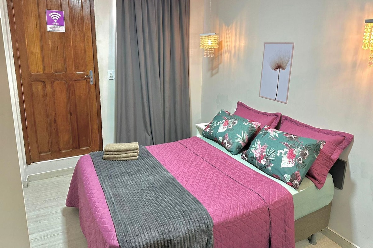 A hotel room with a wooden door, a bed with floral pillows, a grey blanket, and a wall-mounted light fixture.