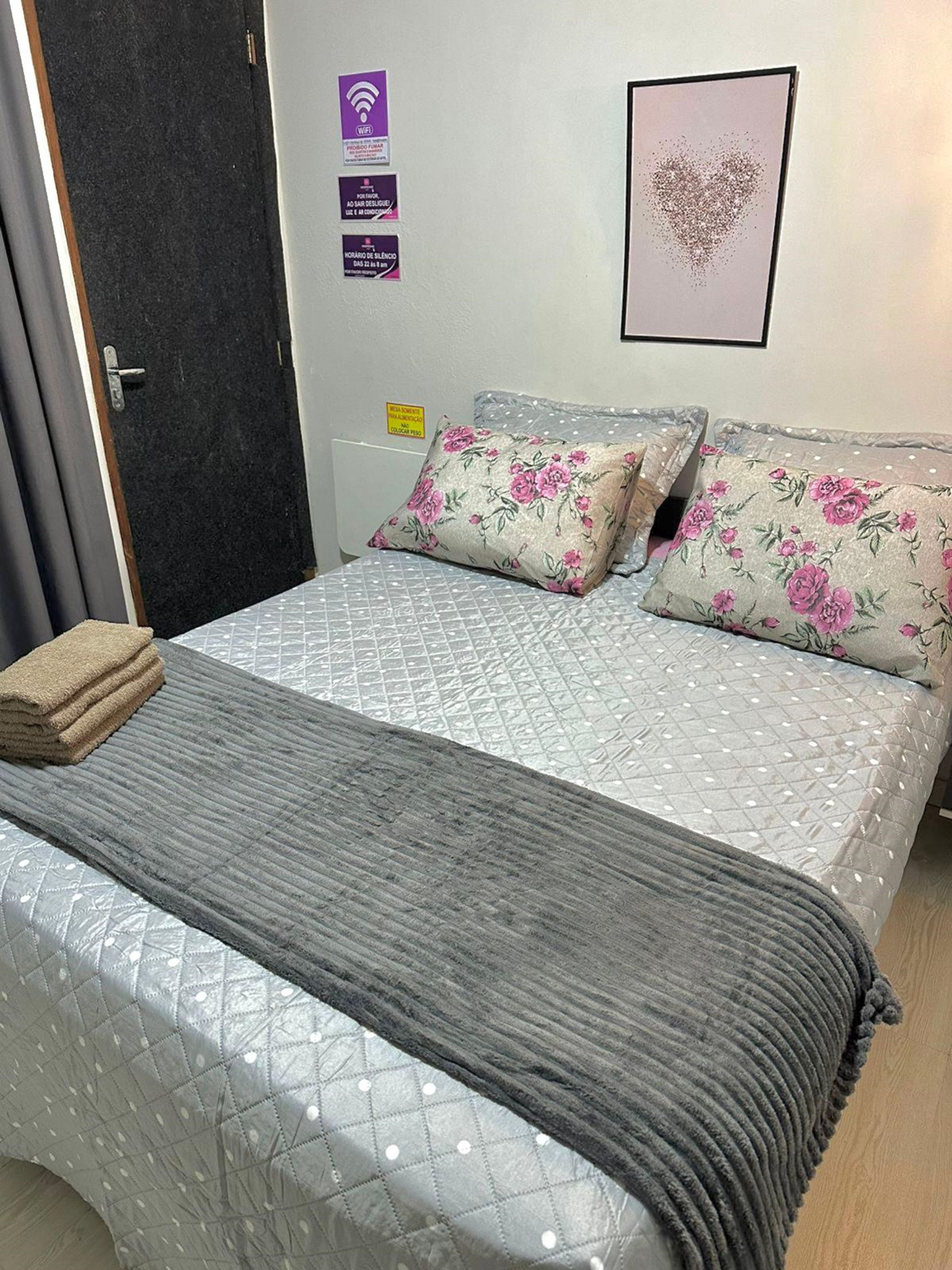 Cama bem arrumada com colcha cinza, duas almofadas florais e manta cinza.