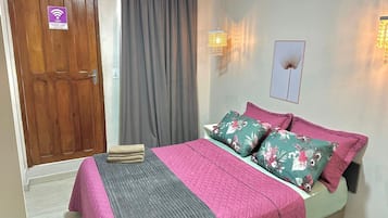 Quarto de hotel com porta de madeira, cama com almofadas florais, manta cinzenta e luminária fixa na parede.