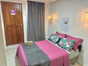 A hotel room with a wooden door, a bed with floral pillows, a grey blanket, and a wall-mounted light fixture. - Hotel Manaus - Dom Pedro (Manaus)