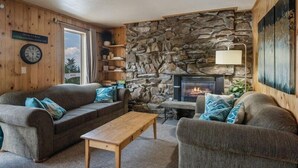 A cozy living room with a stone wall, a fireplace, and wooden furniture. - Der Kastle Ski Condo · Schweitzer Retreat Comfort for all Prime location (Sandpoint)
