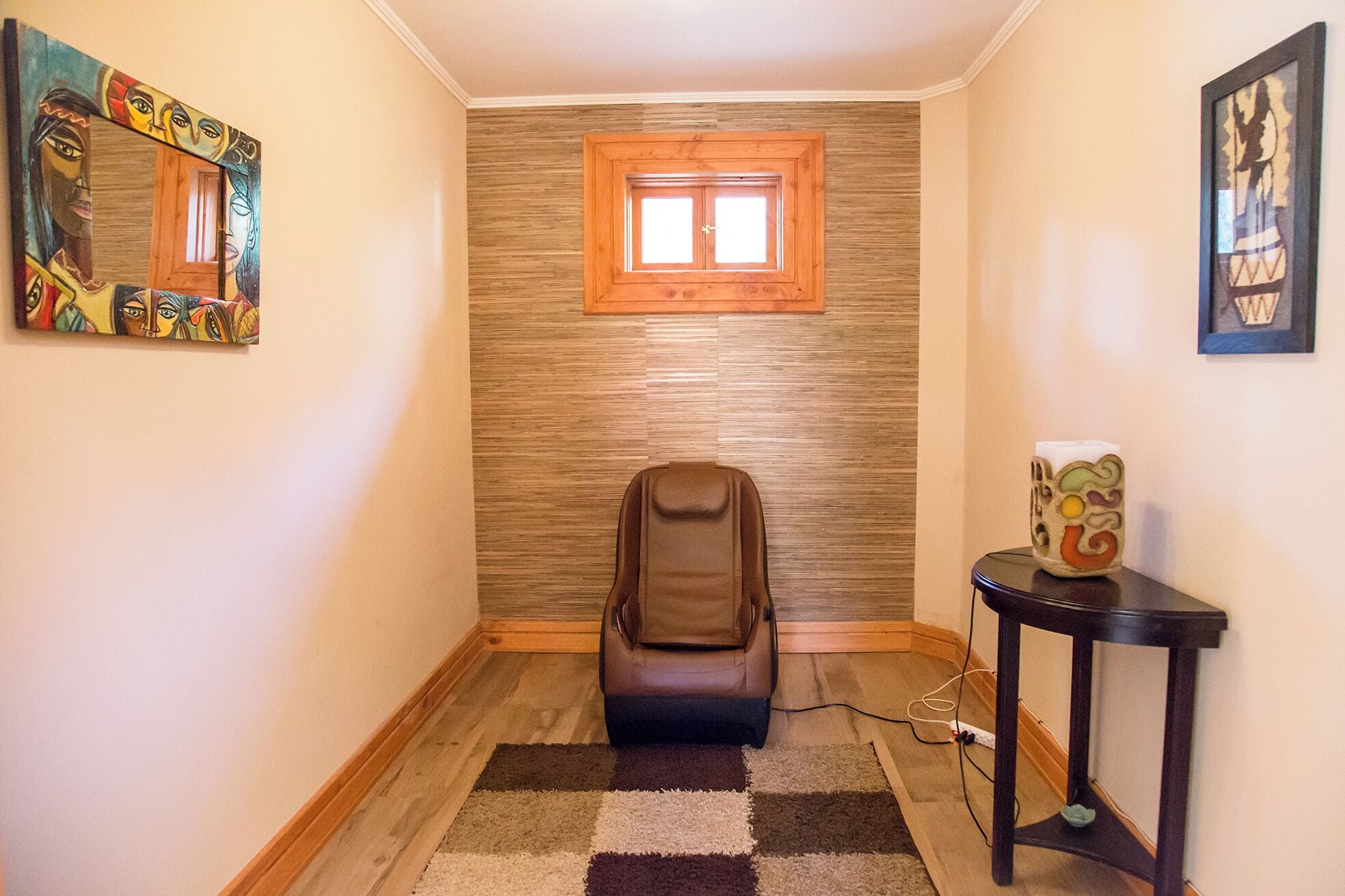 Un pasillo estrecho con una silla de masaje, una mesita con un objeto decorativo y una ventana con marco de madera.