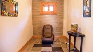 Un pasillo estrecho con una silla de masaje, una mesita con un objeto decorativo y una ventana con marco de madera.