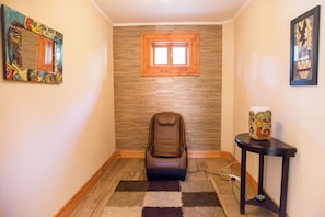 Un pasillo estrecho con una silla de masaje, una mesita con un objeto decorativo y una ventana con marco de madera.