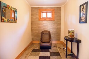 Un pasillo estrecho con una silla de masaje, una mesita con un objeto decorativo y una ventana con marco de madera.