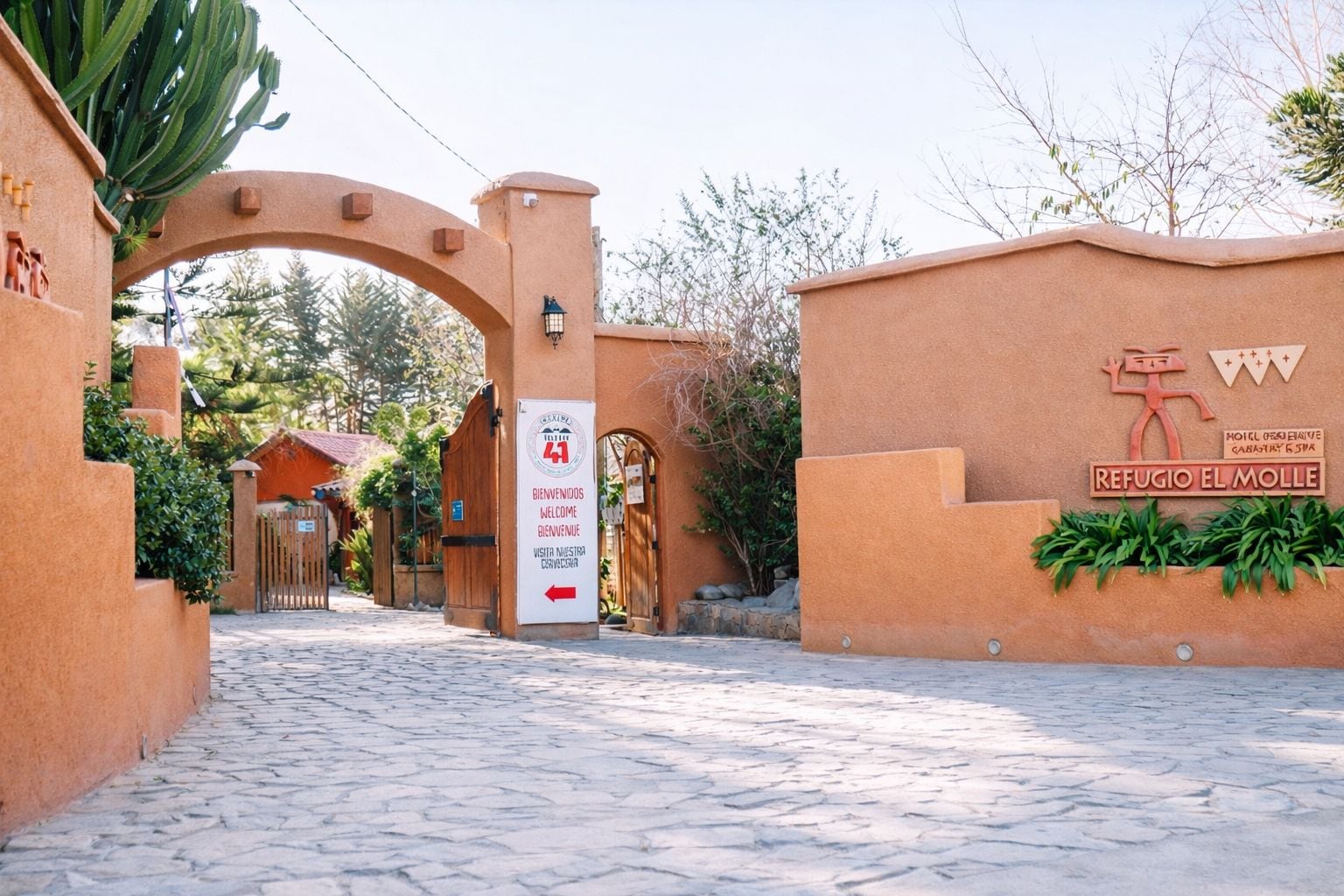 A hotel entrance with a stone pathway leading to a wooden gate. There is a sign indicating the hotel name 'Refugio El Molle' and a welcome message.