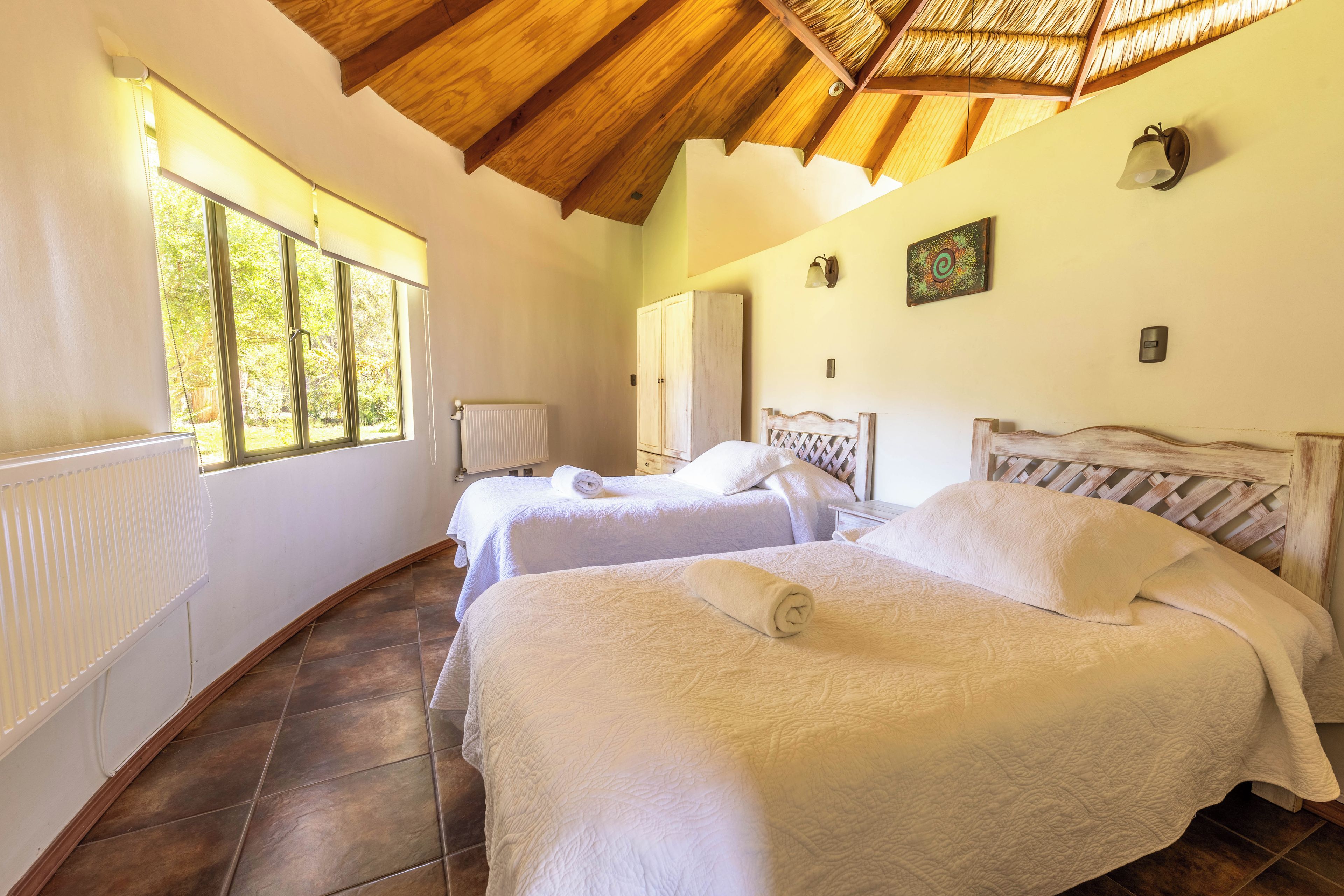 A room with two beds, a radiator, a window, and a ceiling with wooden beams.