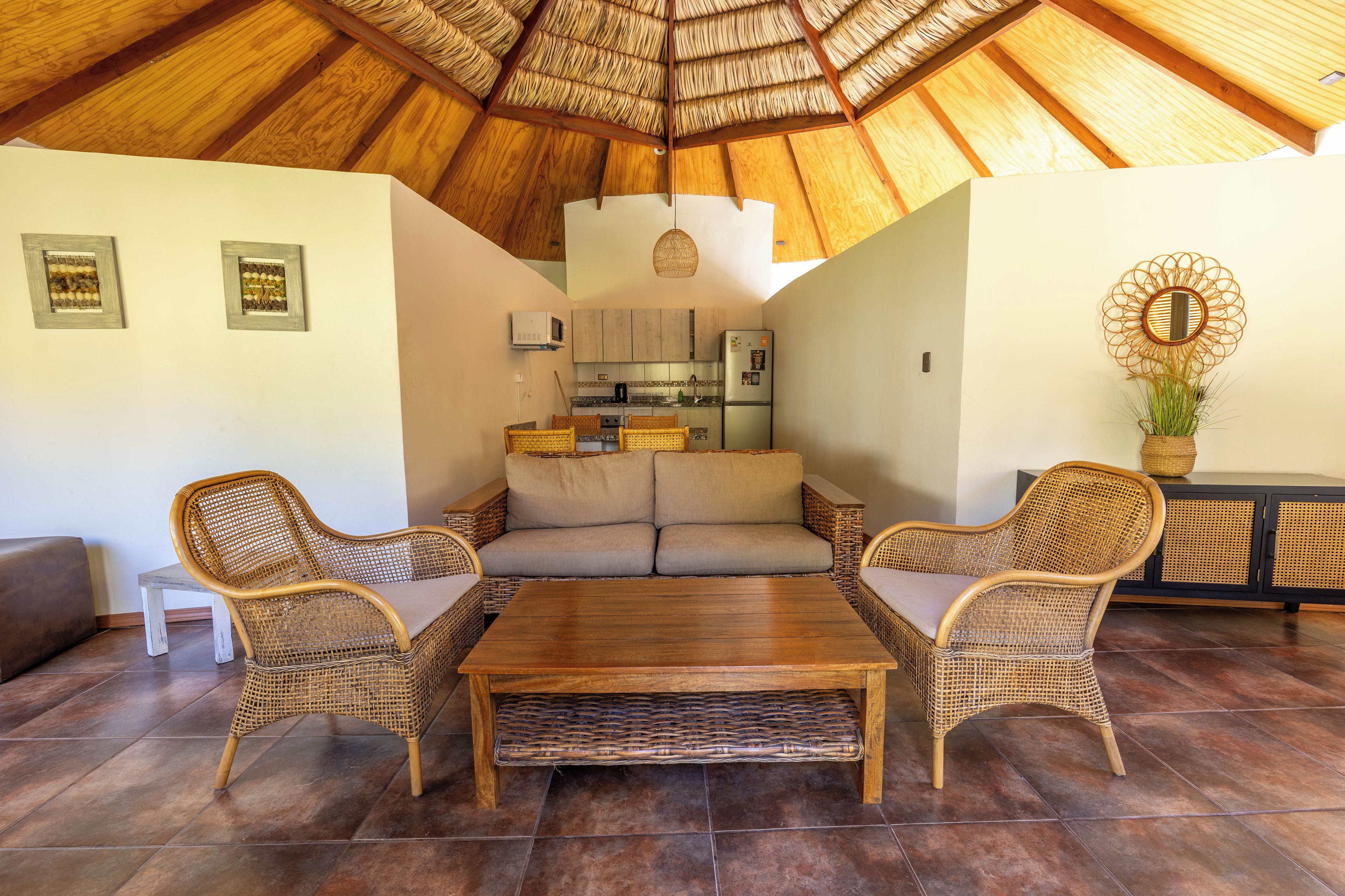 A cozy living area with a thatched ceiling, wicker furniture, and a kitchen in the background.