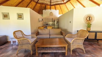 A cozy living area with a thatched ceiling, wicker furniture, and a kitchen in the background.