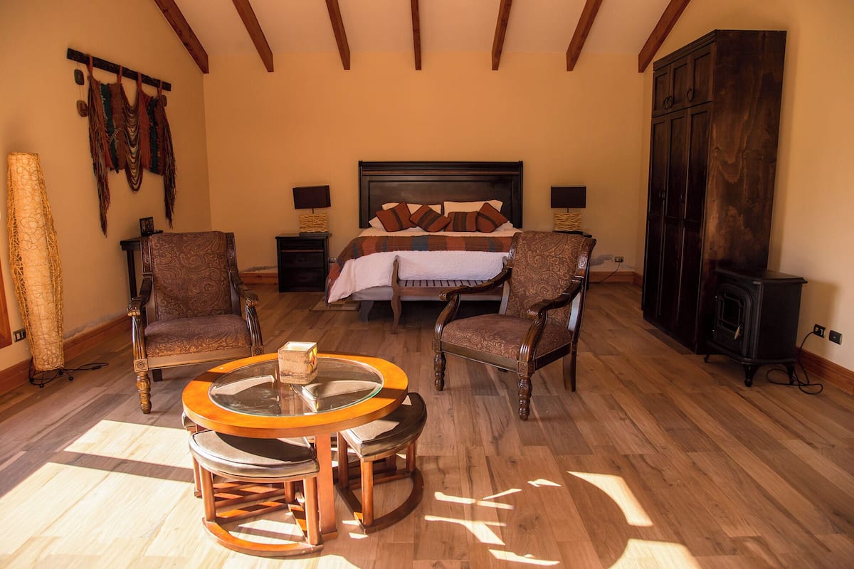 A bedroom with a large bed, two armchairs, a round table, and a wooden cabinet.