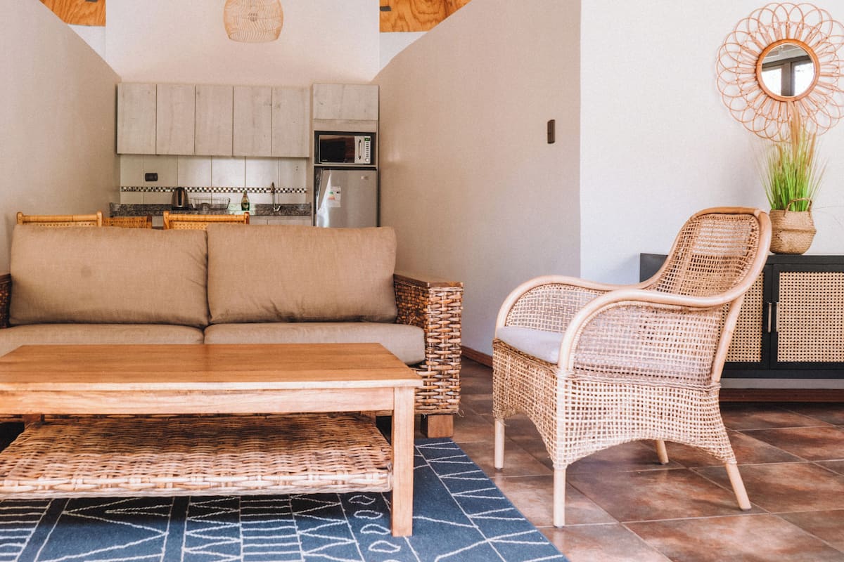 A living room with a sofa, a wooden coffee table, a wicker chair, and a kitchen in the background.