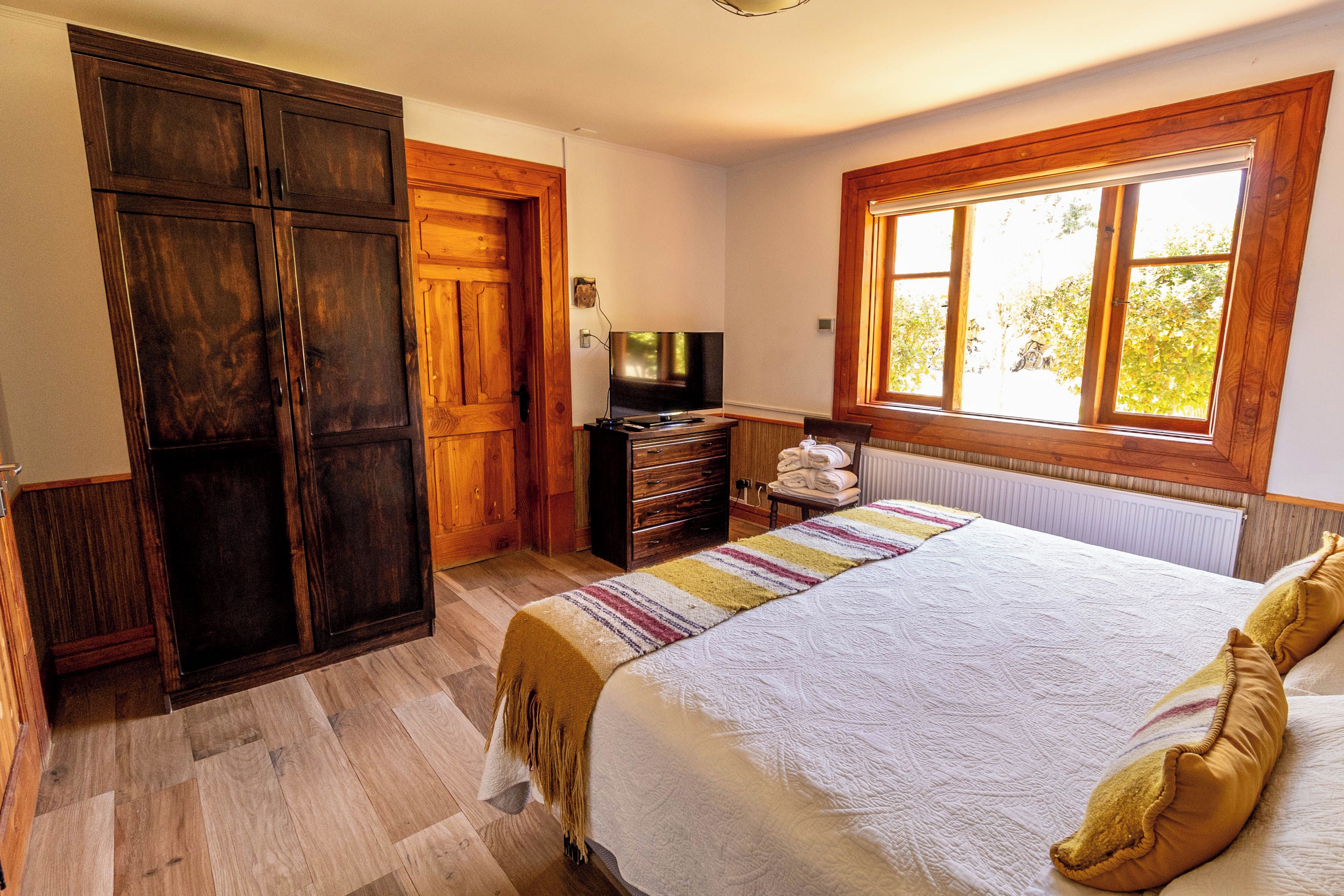 A bedroom with a large bed, wooden wardrobe, a television, and a window with a view of trees.