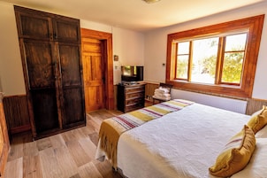 Un dormitorio con una cama grande, un armario de madera, televisión y ventana con vistas a árboles.