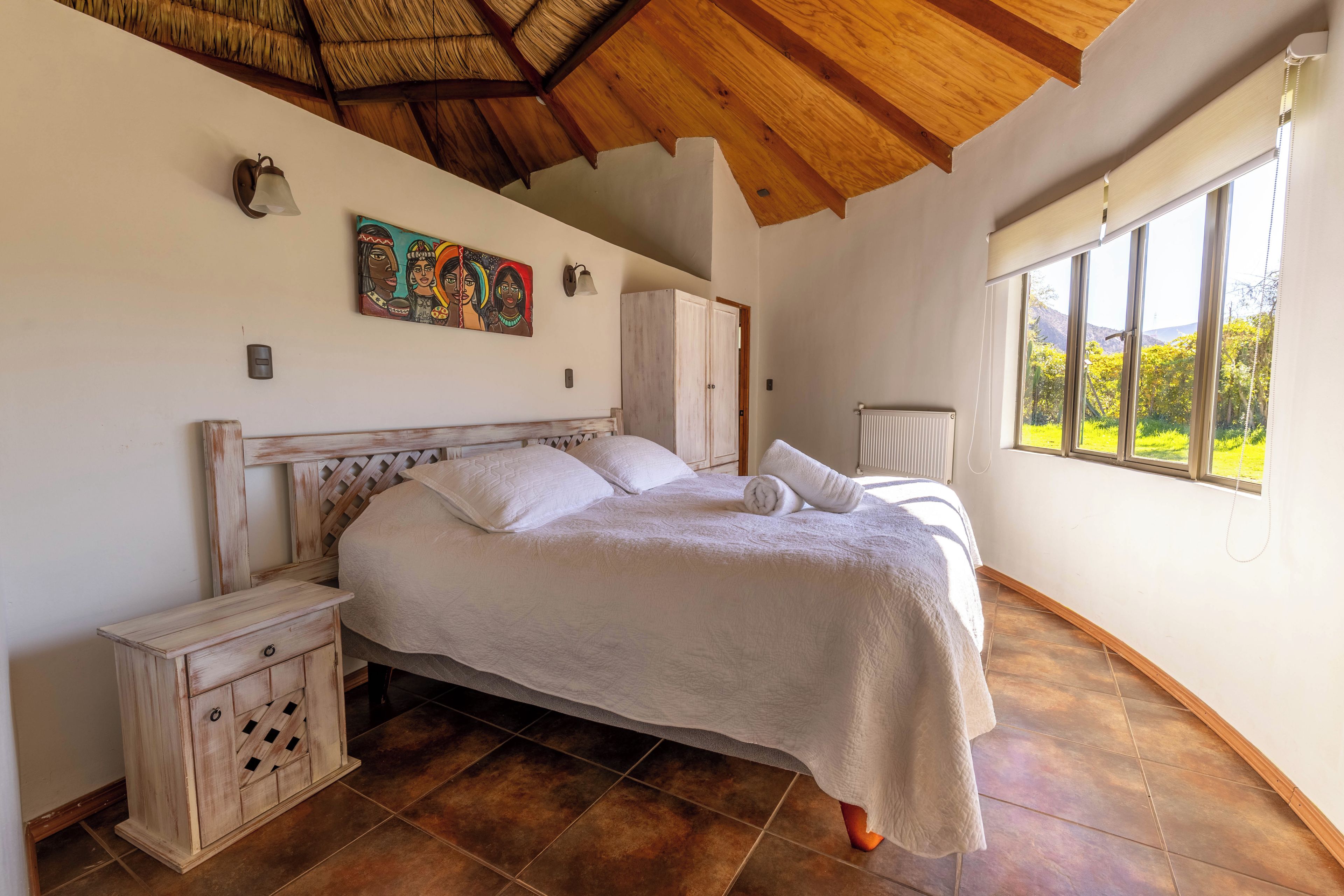 A bedroom with a large bed, a nightstand, a wooden cabinet, a painting on the wall, and a window with a view of greenery.