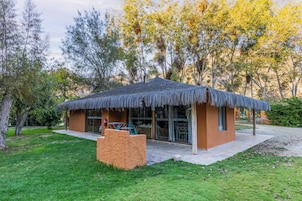 Edificio con techo de paja, puerta y ventanas de vidrio, rodeado de árboles y un área verde.