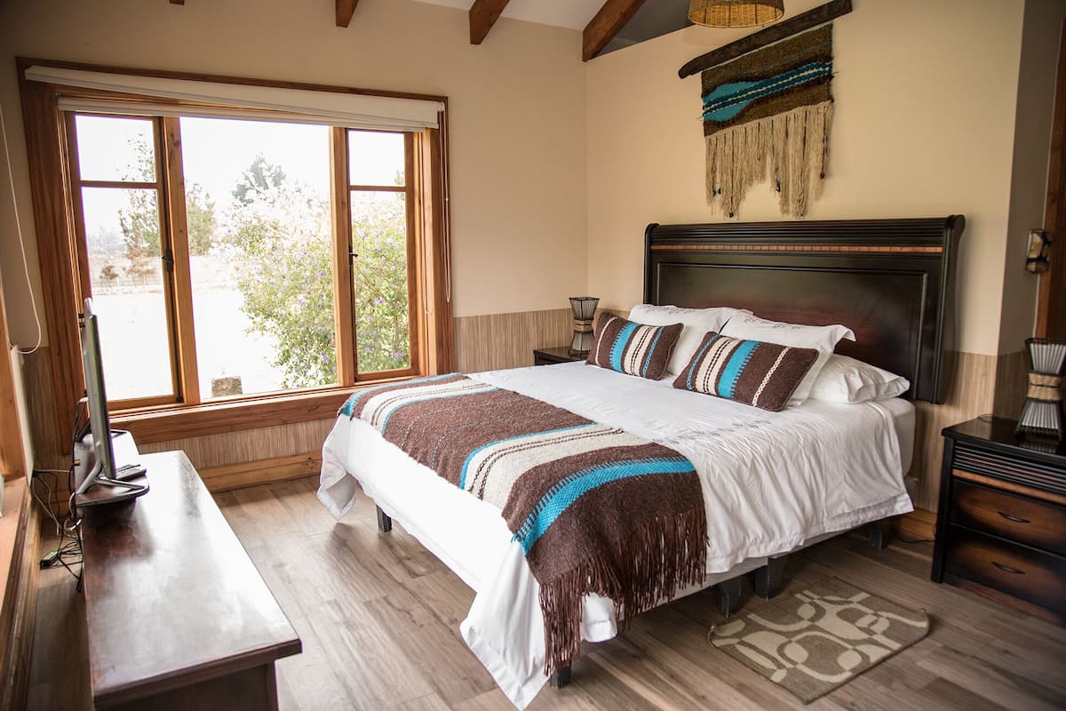 A bedroom with a large bed, wooden headboard, a desk, and a window with a view of trees.