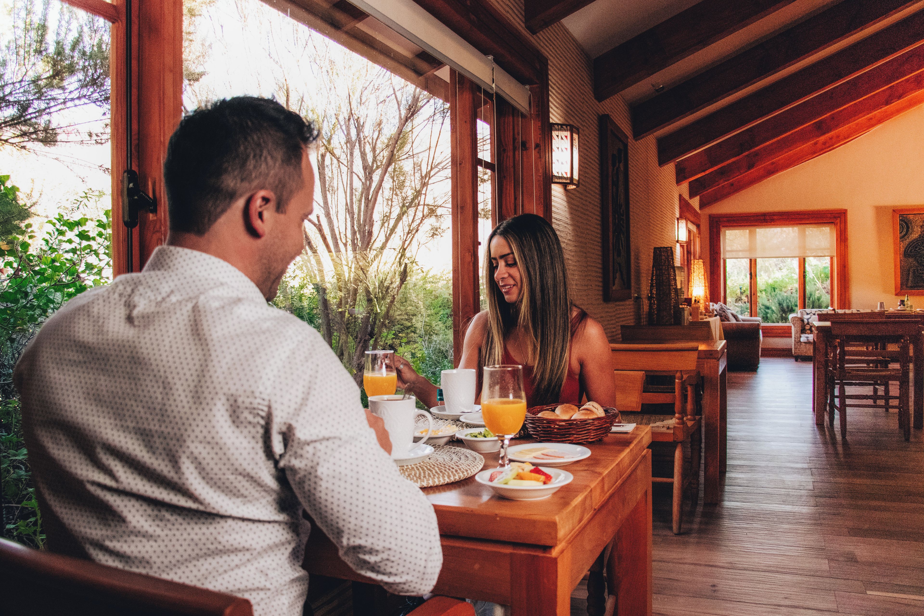 Una mesa de comedor de madera con alimentos para el desayuno, una ventana con vistas a árboles y un techo de madera.