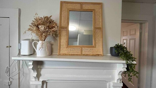 Une cheminée blanche ornée d’un miroir, un vase blanc contenant des fleurs séchées et une petite plante en pot.