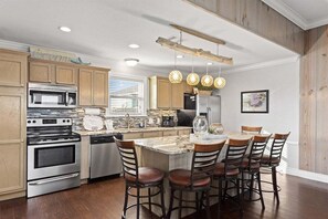A modern kitchen with wooden cabinets, stainless steel appliances, a central island with bar stools, pendant lighting, and a view of the outdoors.