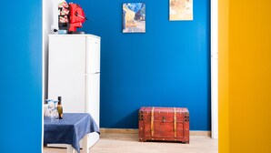 Ein Zimmer mit einem weißen Kühlschrank, einem Tisch mit blauer Tischdecke, einem roten Koffer und zwei Bildern an der Wand.