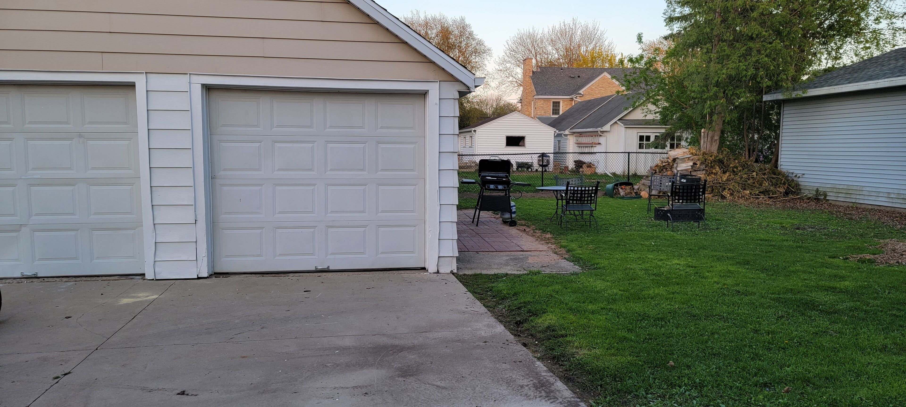 A residential garage with a concrete driveway and a small lawn area with a grill and outdoor furniture.