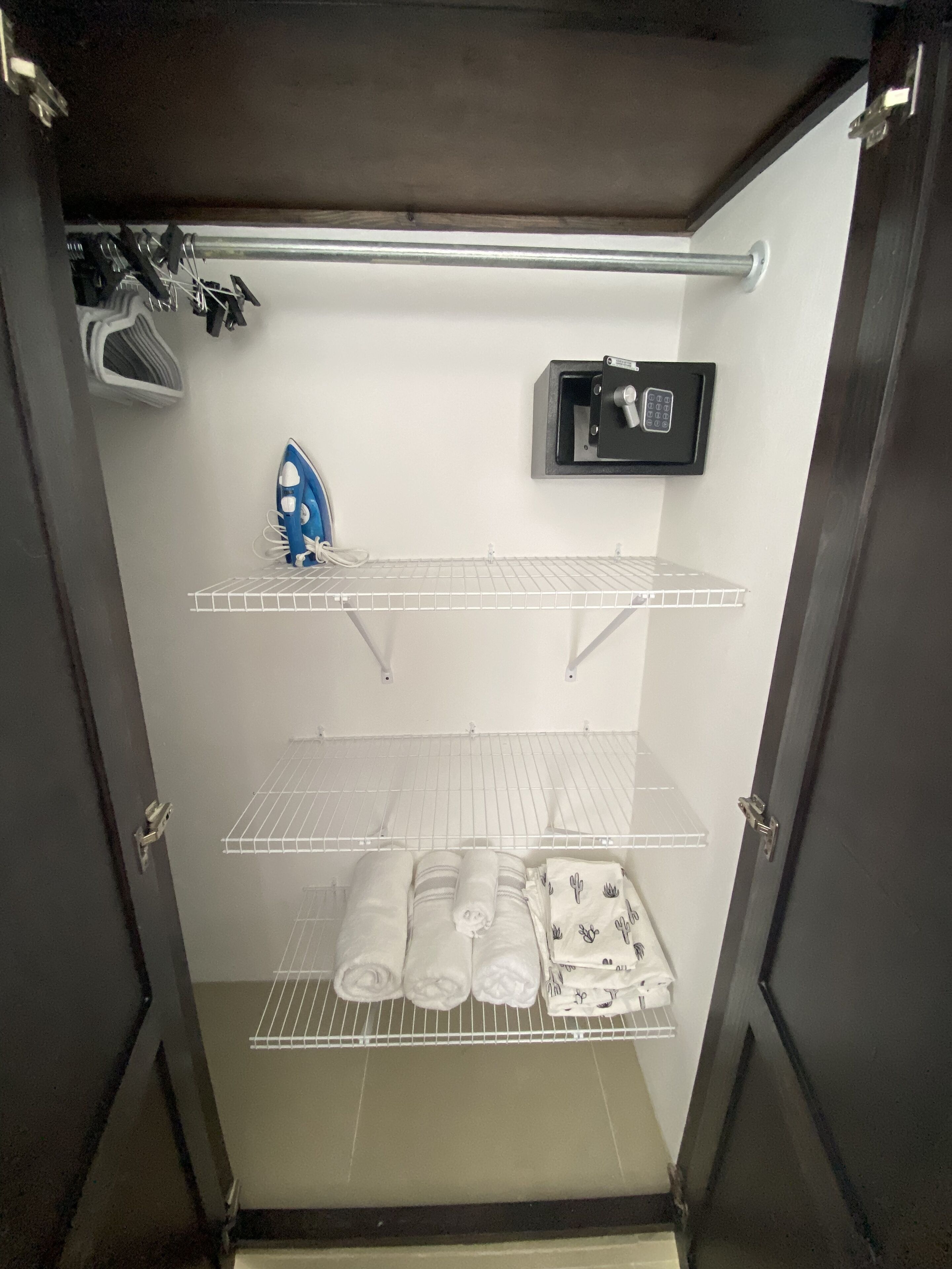 Un armario amplio con estanterías metálicas, una plancha y un panel de control de seguridad.