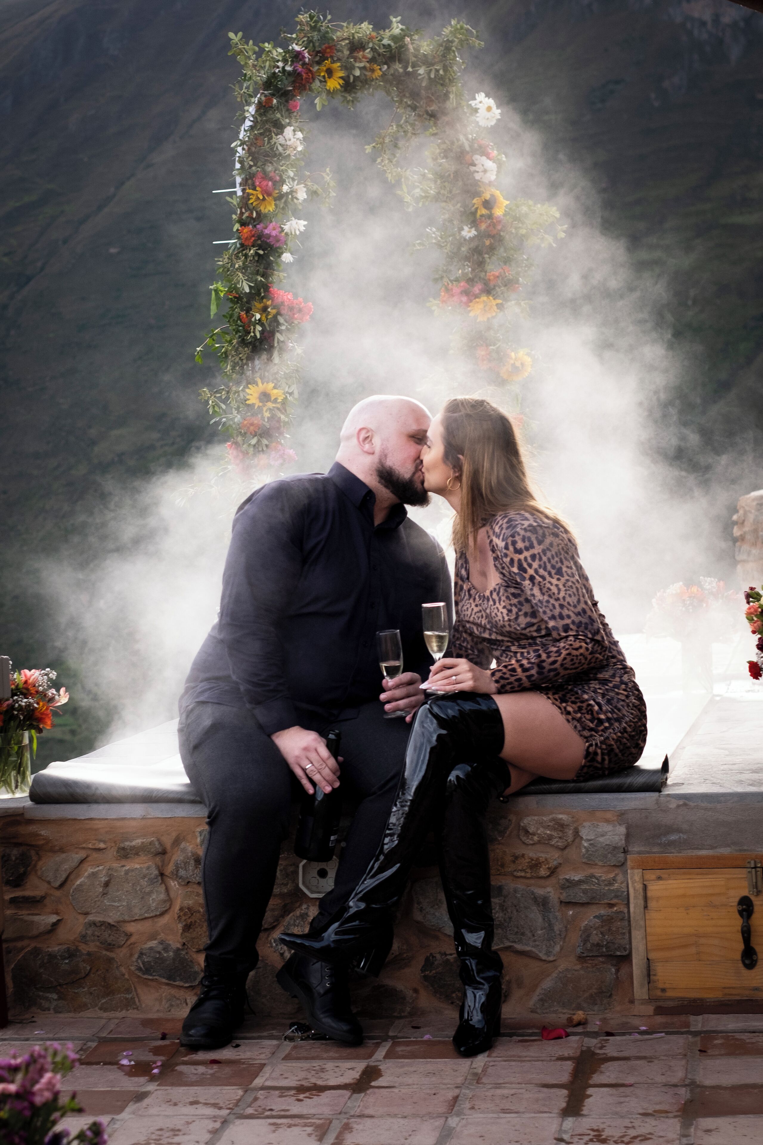 A couple sitting on a stone bench outdoors, surrounded by a floral arch and mist.