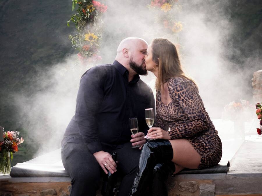 A couple sitting on a stone bench outdoors, surrounded by a floral arch and mist.
