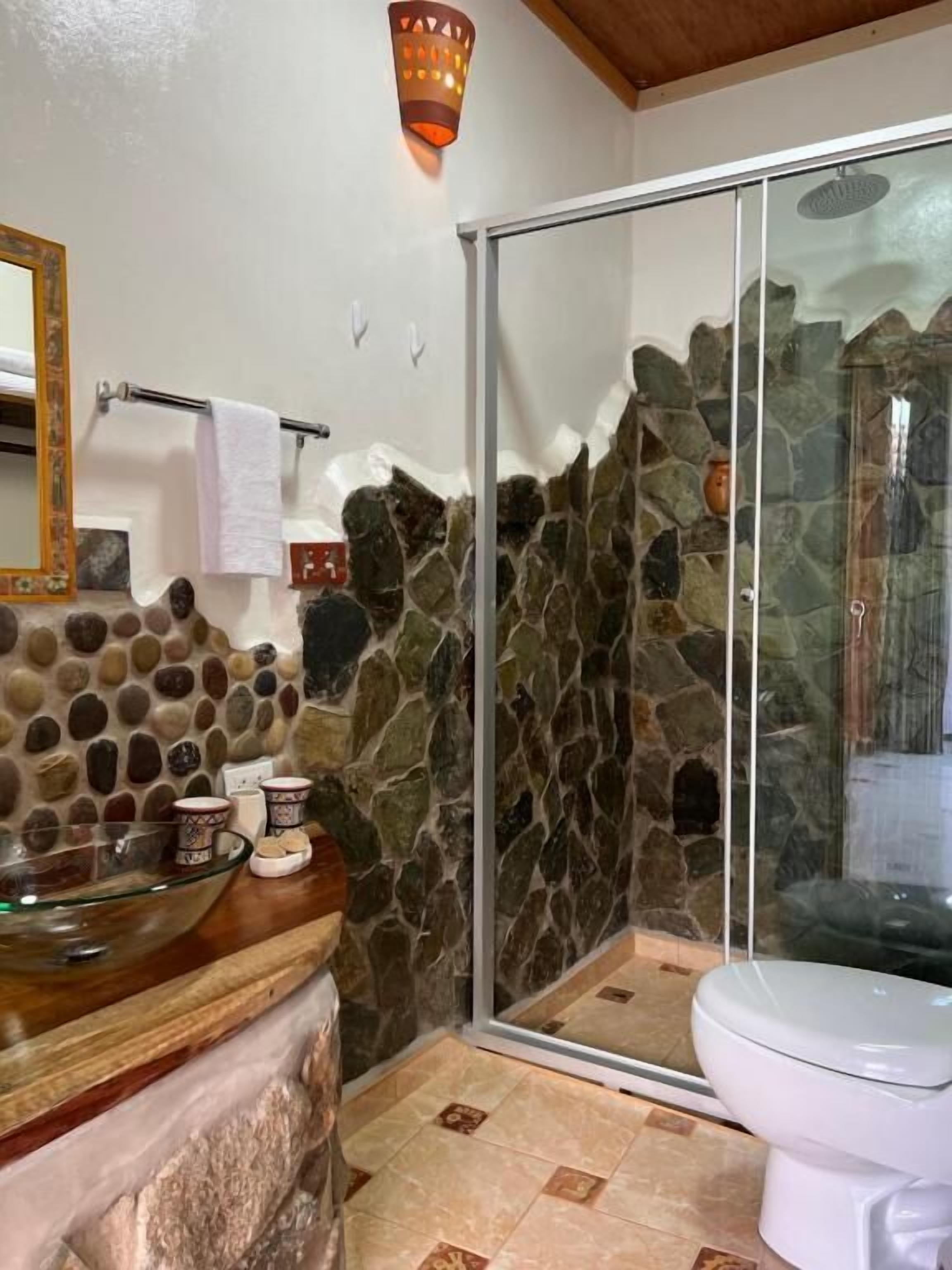 Et badeværelse med stenvæg, brusekabine i glas, glasvask og toilet.