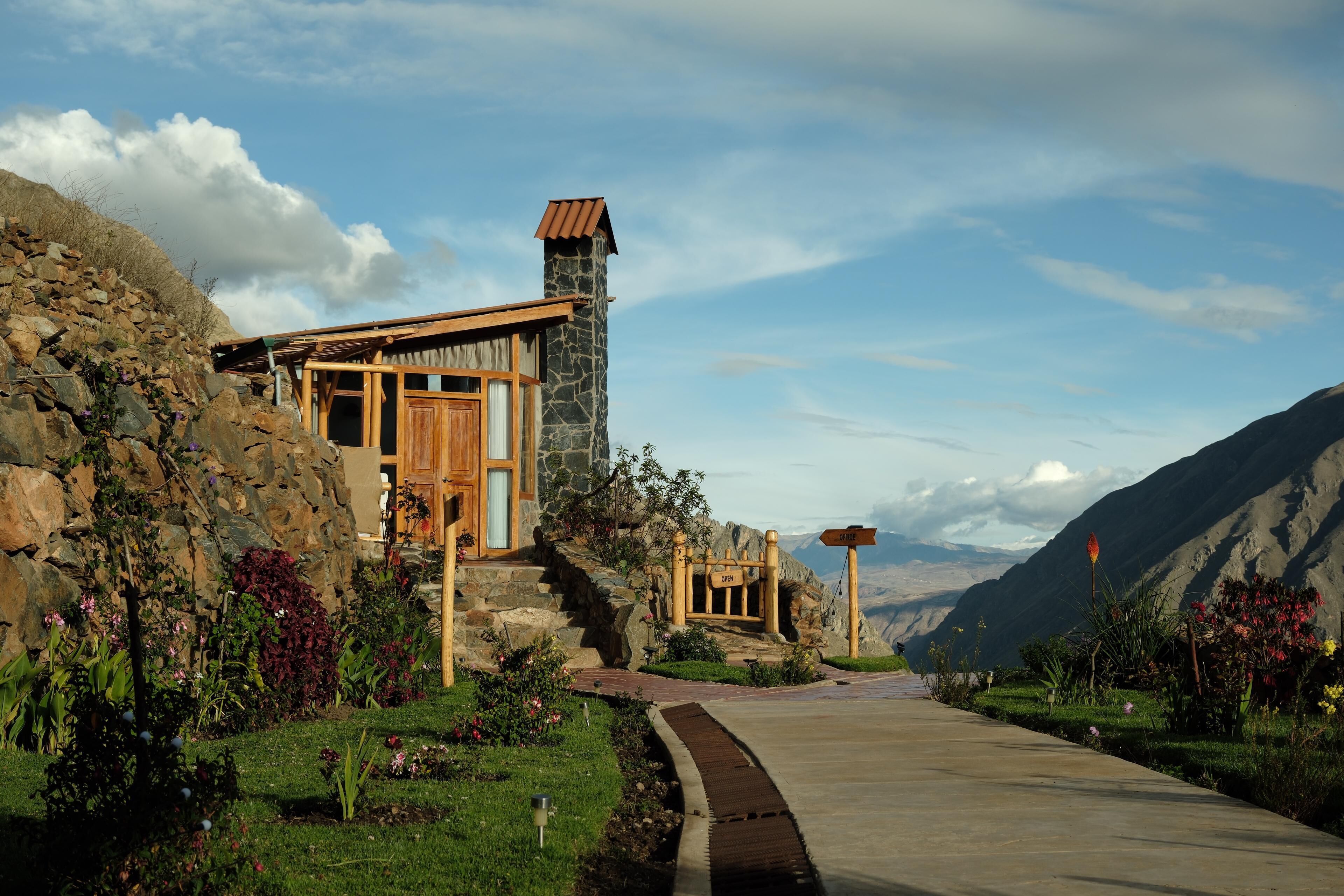 A rustic mountain lodge with a stone chimney, wooden exterior, and a paved driveway leading to a wooden deck.