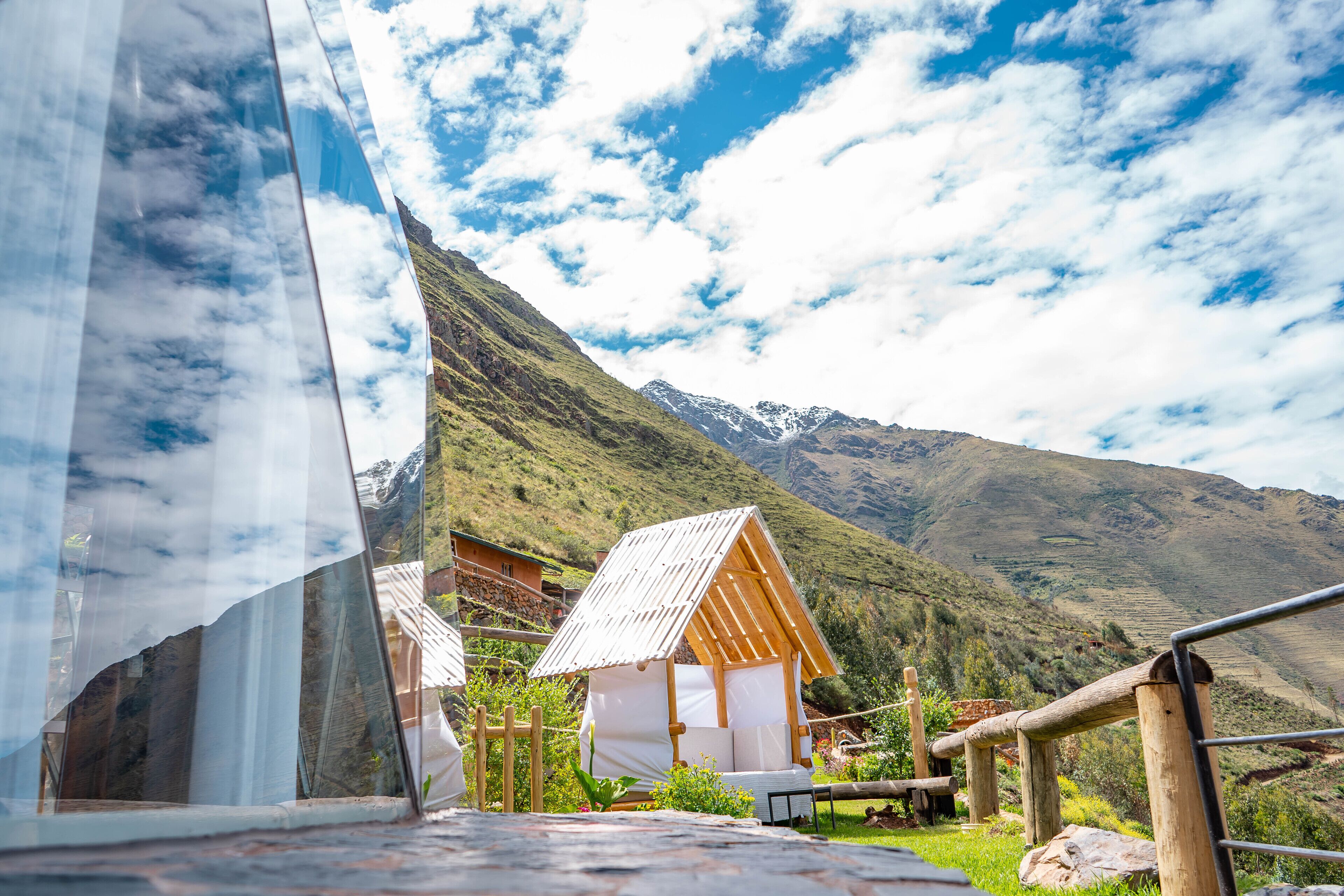 A mountain cabin with a wooden structure and a white roof, surrounded by lush greenery and mountains in the background.
