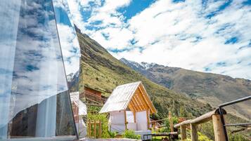 A mountain cabin with a wooden structure and a white roof, surrounded by lush greenery and mountains in the background.
