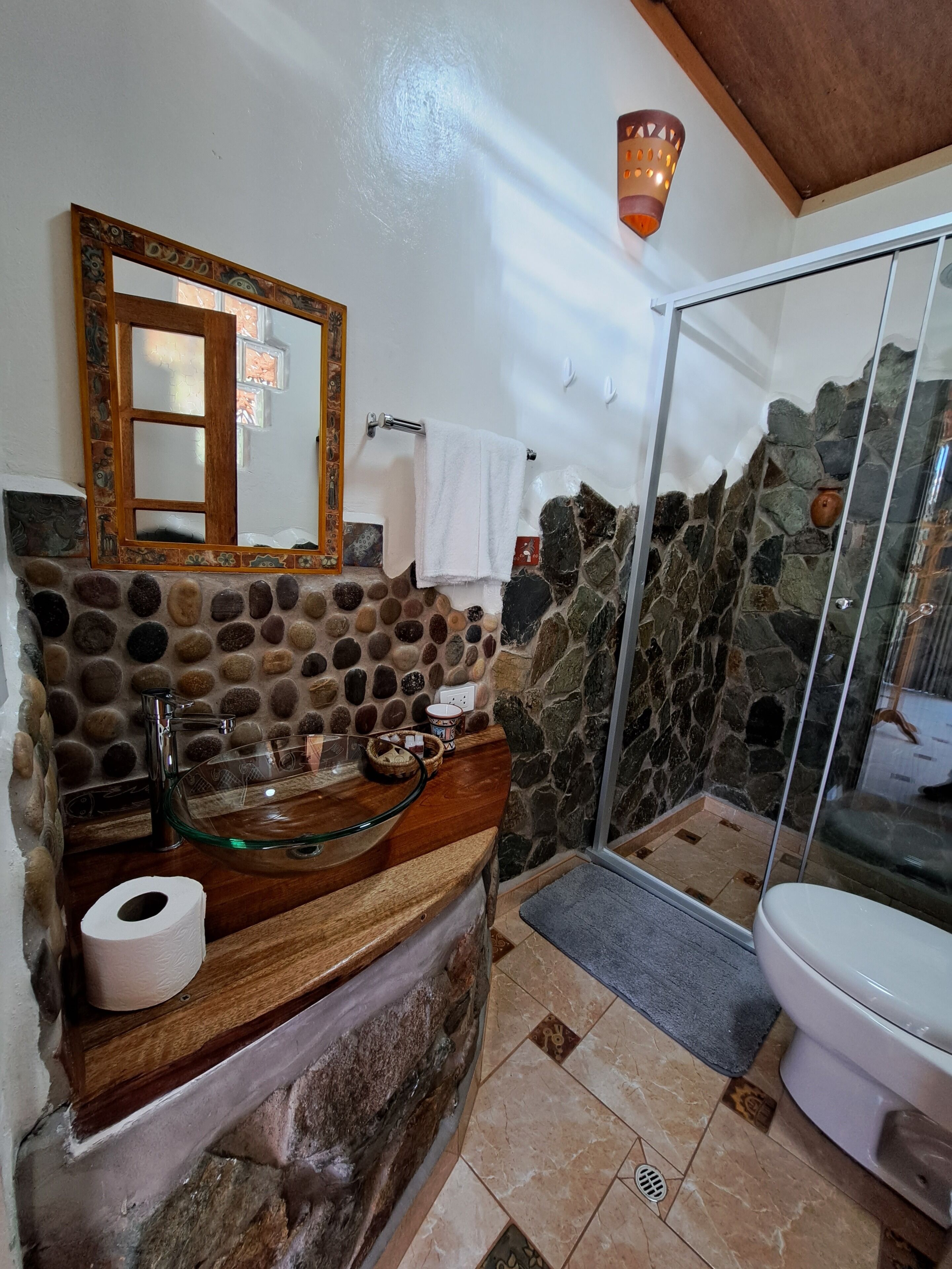 Un baño con pared de piedra, lavamanos de cristal, tocador de madera, espejo y cabina de ducha de cristal.
