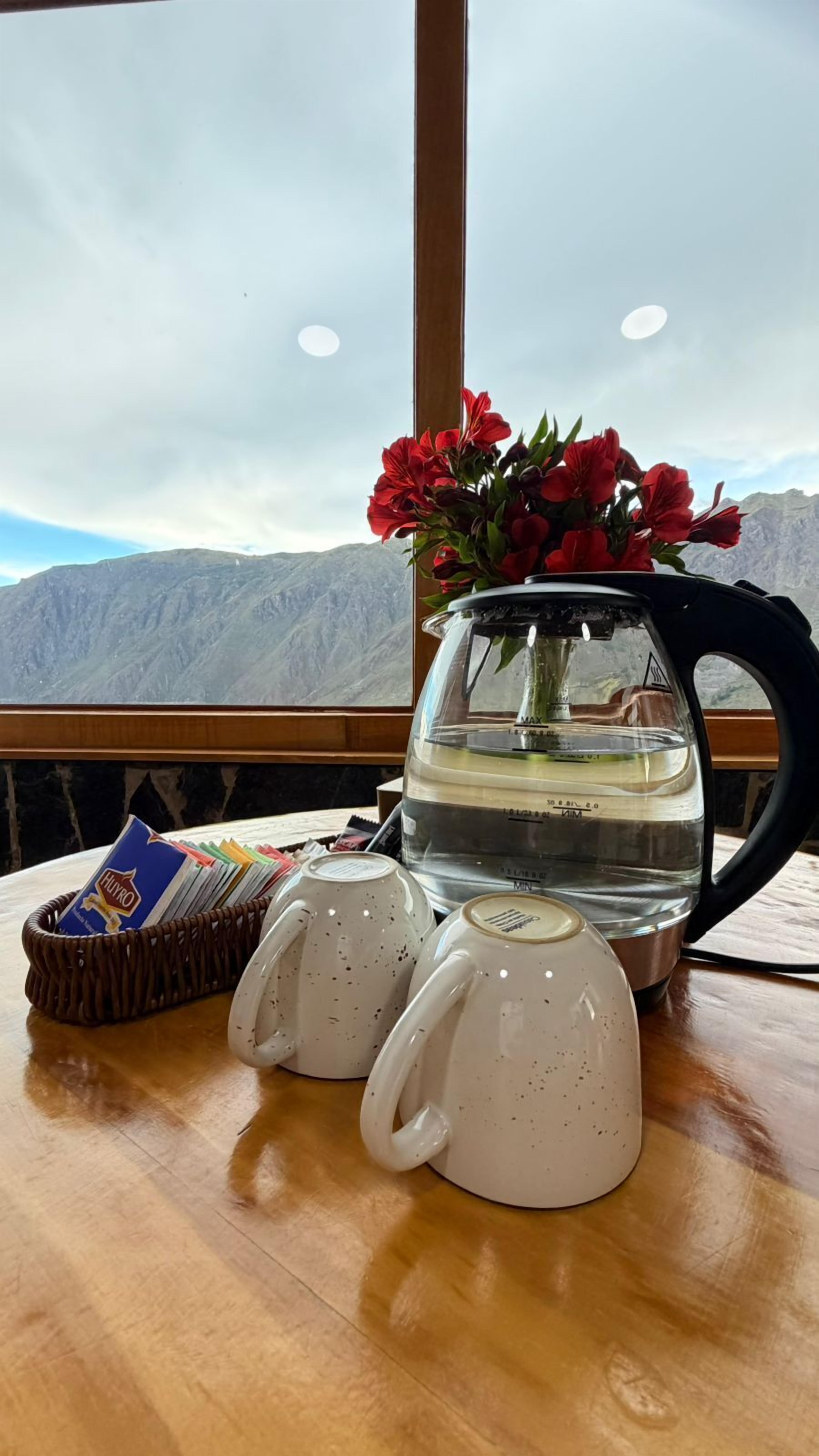 A table with a water kettle, two mugs, a basket of cards, and a vase with red flowers.