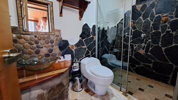 A bathroom with a stone wall, a glass shower enclosure, a wooden vanity, and a glass sink.