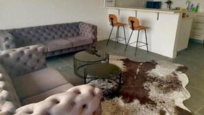 Una sala moderna con un sofá capitonado, una mesa de centro redonda y un tapiz de cuero de vaca. Hay dos taburetes de bar y una cocina con gabinetes blancos y una encimera.
