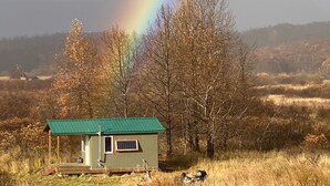 A small cabin with a green roof, surrounded by trees and a grassy field, with a rainbow in the background. - Saltery Adventures Remote Loft Cabin (Kodiak)