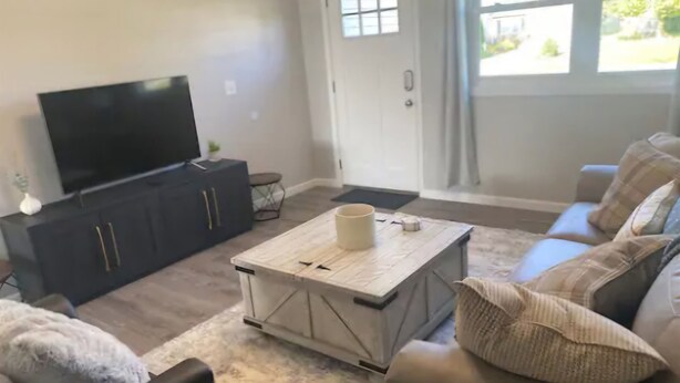 Ein Wohnzimmer mit Flachbildfernseher auf einem schwarzen Schrank, einem Couchtisch, einer Couch mit Kissen und einer Tür mit Blick ins Freie.