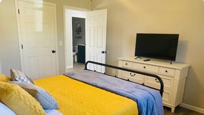 Ein Schlafzimmer mit einem Bett, einem Schminktisch, einem Fernseher und einer Tür zu einem anderen Raum.