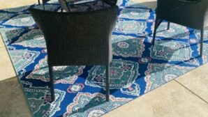 Ein überdachter Patio-Bereich mit einem gemusterten Teppich, Gartenmöbeln und Blick auf einen Hinterhof.