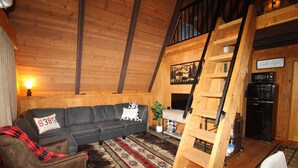 Ein gemütliches Blockhaus-Interieur mit Holzbalkendecke, einer schwarzen Ledersofa, einer Holzleiter und einer Küchenzeile.