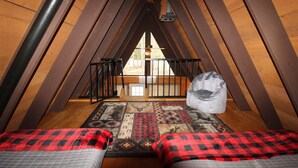 Ein gemütliches Schlafzimmer im Dachgeschoss mit schrägen Wänden, einem Bett mit einer rot-schwarz karierten Decke, einem gemusterten Teppich und einem großen Sack auf dem Boden.