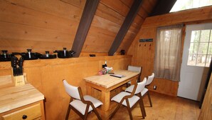 Ein Holzblockhaus-Interieur mit einem Esstisch, vier Stühlen, einer Küchenzeile mit Töpfen und Besteck sowie einem Fenster mit Blick auf Bäume.