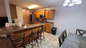 A kitchen with wooden cabinets, a central island with bar stools, a refrigerator, microwave, and a trash can. - Heart of Branson #2 (Branson)