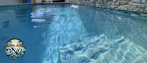 A swimming pool with clear water, a stone wall, and a covered seating area.