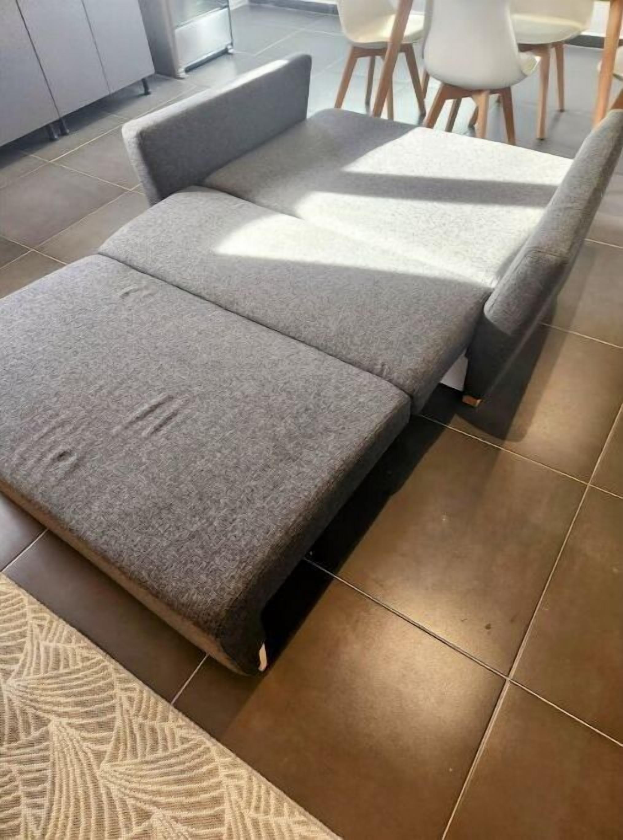 Um sofá moderno com tecido cinza, posicionado sobre um chão de azulejos com um tapete estampado por baixo.