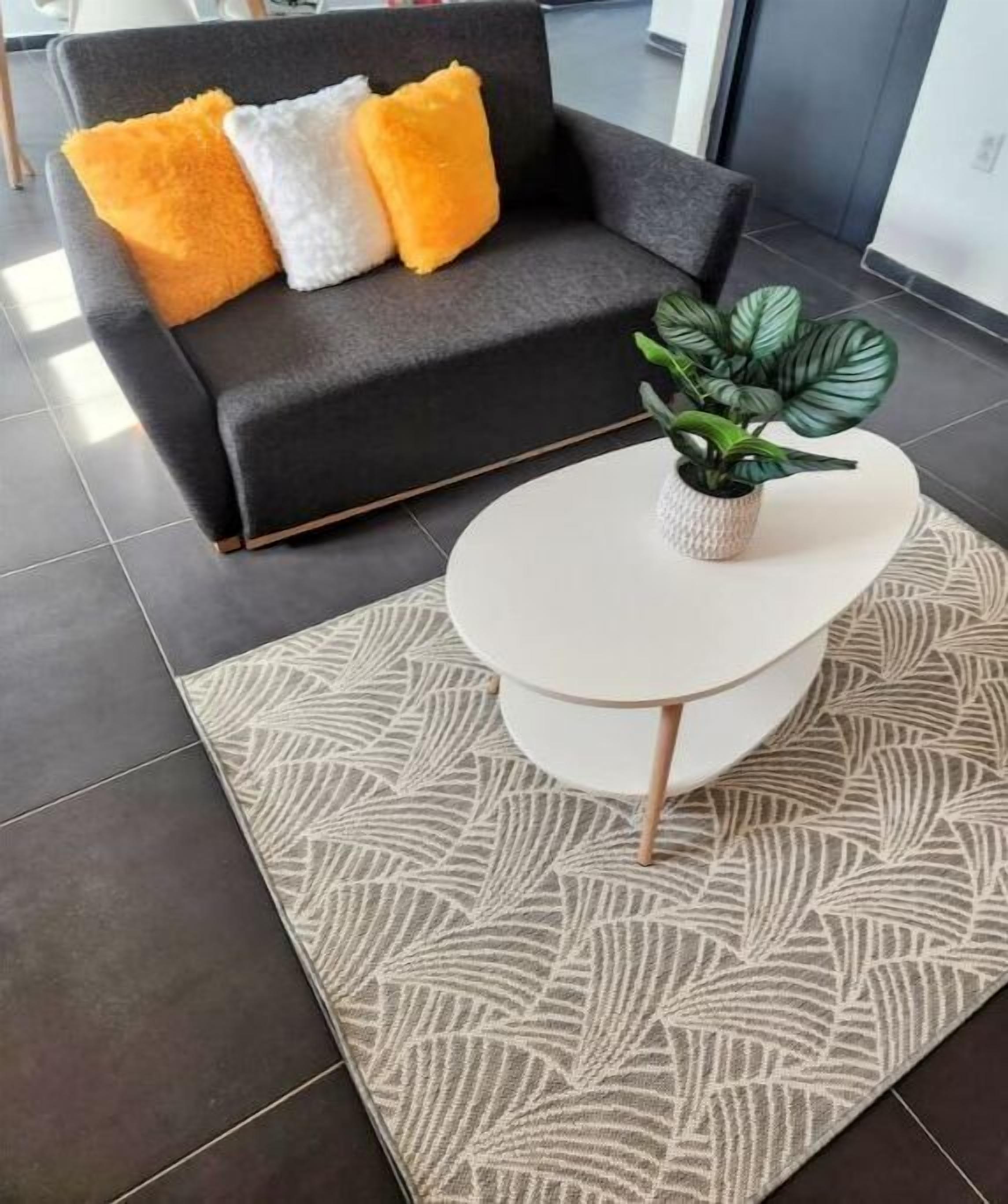 Ein modernes Wohnzimmer mit einem dunkelgrauen Sofa, drei Kissen in Orange, Weiß und Gelb sowie einem weißen runden Couchtisch mit einer Pflanze.