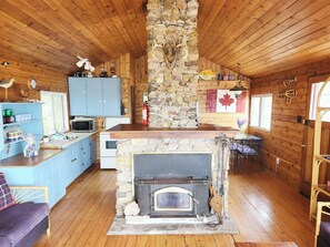 A cabin interior with a stone fireplace, wooden walls, a kitchen area, and a dining space.